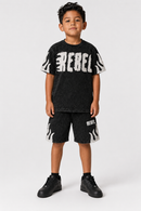 A young boy modeling a black Rebel brand short-sleeve t-shirt and matching black shorts with white flame and logo graphics, paired with black sneakers, against a light gray studio background.