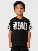 A young boy wearing a black acid-wash t-shirt with a distressed white 'REBEL' graphic across the chest, photographed against a light gray studio background.