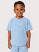 A young boy wearing a light blue t-shirt and matching shorts set, featuring the word 'Saint' in a white Gothic font across the chest and on the shorts, photographed against a plain light gray background.