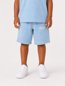 A child wearing light blue denim shorts with gold gothic lettering on the left thigh, styled with a matching light blue t-shirt and white sneakers, photographed against a clean white studio background.