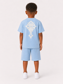 A child model shown from the back wearing a light blue short-sleeve t-shirt and matching shorts, featuring a large white ornate cross graphic on the back, paired with white sneakers, against a plain light background.