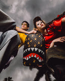 A low-angle lifestyle shot of a person wearing a black and white checkered shark-mouth backpack with red, white, and yellow flame details, posed between two other models against a dark, cloudy sky.