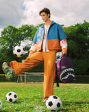 A man stands on a grassy soccer field wearing a color-blocked blue, orange, and white jacket with matching orange pants, kicking a soccer ball while holding a purple shark-mouth backpack.