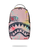 A Sprayground Shark backpack in pink, featuring a graphic print of a shark mouth and colorful paint splatters, with a top handle and zippered compartments.