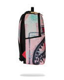 A Sprayground shark backpack in pink, black, and turquoise paint splatter print, shown from the side on a white background with a soft shadow underneath.