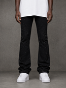 A person wearing black flared jeans with small white speckled details, styled with a white t-shirt and white sneakers, standing against a gray textured studio background.