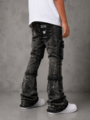 A child wearing black heavily embellished denim flare jeans with sequins and fringe, paired with a white t-shirt and white sneakers, shown from the back against a gray concrete wall.