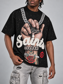 A man wearing a black Saint Laurent graphic t-shirt with a detailed print of a hand, a cross necklace, and a rose, paired with distressed light gray cargo jeans against a solid gray background.