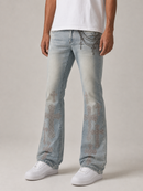 A person wearing light-wash blue jeans with decorative chain and rhinestone embellishments on the front and upper thighs, paired with a white t-shirt and white sneakers, against a neutral studio background.